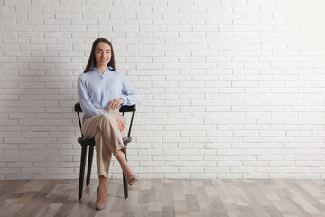 Young businesswoman sitting in office chair near white brick wall indoors, space for text