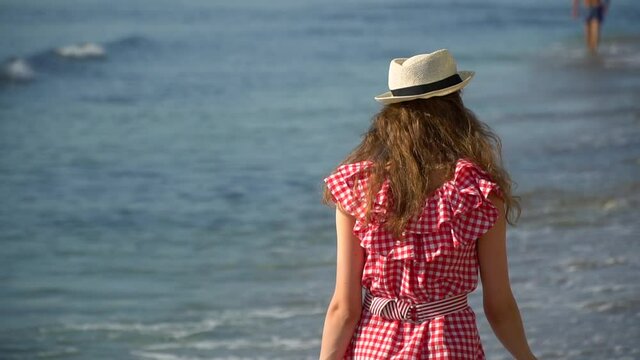 From the back, a shot of a woman in a dress on the beach. An adult girl walks along the beach on a summer sunny day. Enjoy your vacation and relaxation at the resort.