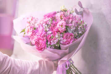 A hand holds a bouquet of light pink, purple, white cute delicate small roses of different sizes, flowers of green leaves. Paper packaging.