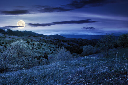 rural mountain landscape in spring at night. grass and trees on hills rolling through the green valley in to the distant ridge beneath a cloudy sky in full moon light