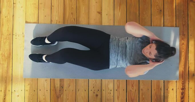 Athletic lady in tracksuit and sneakers does crunches lying on grey mat put on wooden floor at home upper view slow motion