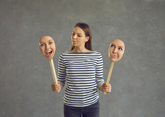 Woman stands on a gray background and holds two faces with different emotions and moods. Brunette...