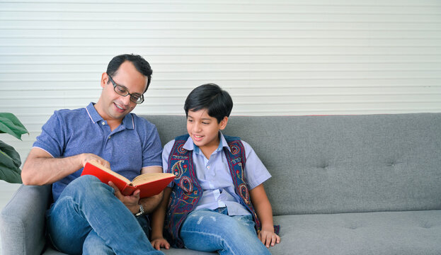 Happy indian father reading book with his son in living room.