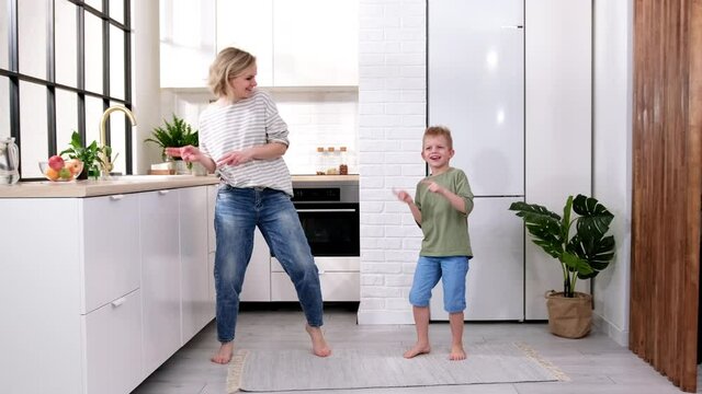 Excited mom or babysiter and kid boy jumping, dancing, laughing in modern scandinavian house kitchen. Happy family mother and son having fun enjoy playing singing together at home
