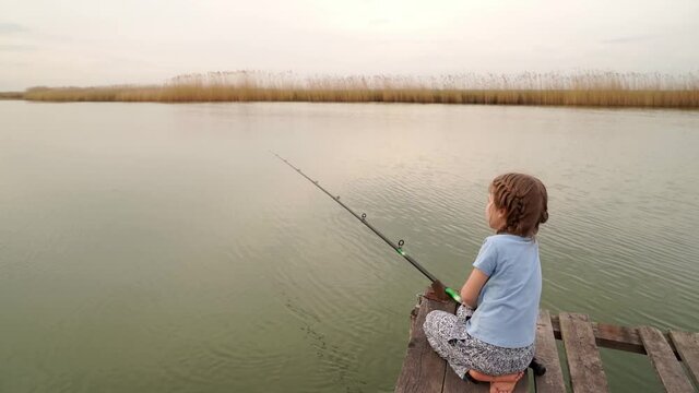 a little girl sits with a fishing rod on a bridge by the river. 