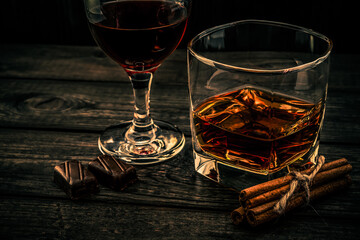 Glass of brandy and red wine with chocolate and cinnamon sticks tied with jute rope on an old wooden table. Angle view, focus on the cinnamon sticks