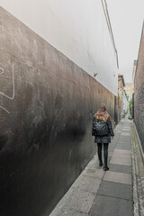 Mujer joven de pelo largo vestida de negro y cargando una mochila, caminando hacia el final de una calle estrecha con muros de ladrillos - obrazy, fototapety, plakaty