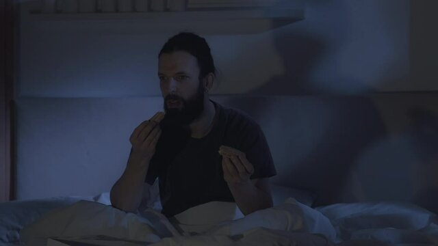 Night eating. Food coaching. Healthy lifestyle. Curious man watching nutrition TV program disgusted with sweet pastry in bed late in dark bedroom.
