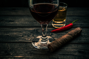 Glass of red wine and tequila with cuban cigar and cayenne pepper on an old wooden table. Close up view, focus on the cuban cigar