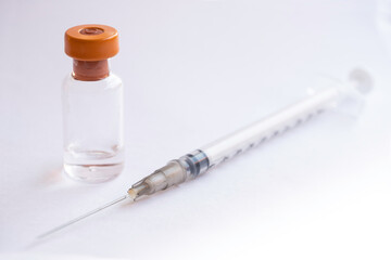 Thin syringe in front of a single corona vaccination vial with orange cap on a white background. Focus on the orange cap of the bottle, shallow depth of field