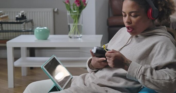 Side view of bored sad African American woman printing Remote working on label maker sitting on bag chair with tablet. Young manager tired of home office. Cinema 4k ProRes HQ
