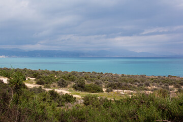 Landscape of Cabo de las Huertas, in Alicante, with vegetation in the foreground, the calm Mediterranean Sea and mountains on the horizon on a cloudy day.