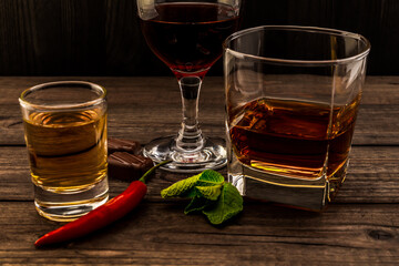 Three glasses with brandy, tequila and red wine with mint sprig, chocolate and cayenne pepper on an old wooden table. Close up view