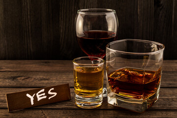 Three glasses with brandy, tequila and red wine with the wooden plank on it is an inscription "Yes" on an old wooden table. Angle view focus on the inscription