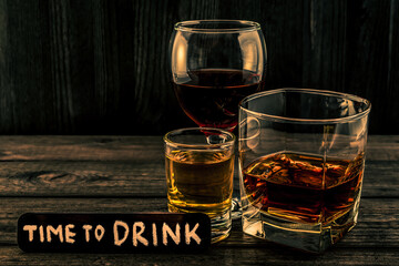 Three glasses with brandy, tequila and red wine with the wooden plank on it is an inscription "time to DRINK" on an old wooden table. Angle view focus on the inscription