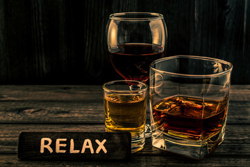Three glasses with brandy, tequila and red wine with the wooden plank on it is an inscription "RELAX" on an old wooden table. Angle view focus on the inscription