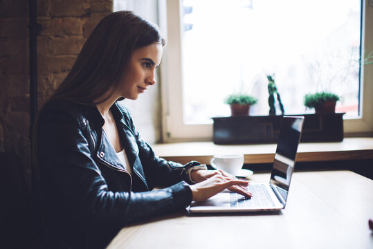 Side view of skilled female blogger typing text for sharing content publication in social media during laptop networking in cafe interior, millennial IT professional programming data software