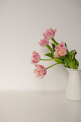 A bouquet of fresh pink parrot tulips with green leaves in a white ceramic jug standing on a table against paper backdrop. Flower decorative composition.