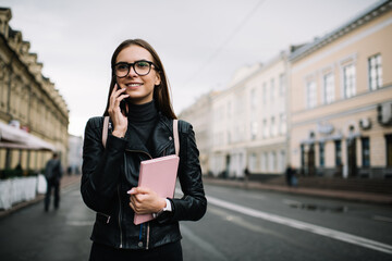 Obraz premium Millennial female student with education textbook using roaming internet connection for phone talking, happy tourist in optical eyewear making international conversation via smartphone application