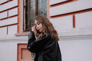 Smiling young woman in black leather jacket standing in front of white wall touching hair, looking over shoulder. Urban style and street fashion. Girl with curly hairstyle in casual outfit