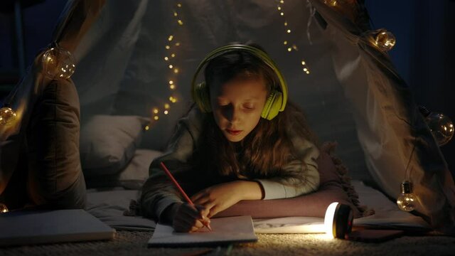 Pretty girl in headphones drawing with colored pencils while lying on floor in beautiful decorative tent at home. Teen singing and listening to music while spending free time in evening.