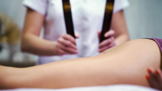 Legs of woman lying on couch and getting bamboo sticks massage. Partial view therapist in white uniform carrying out body care procedures in spa center. Concept of wellness
