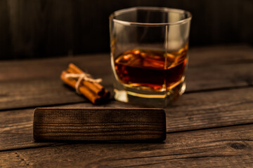 Glass of brandy with cinnamon sticks tied with jute rope and the empty wooden plank on an old wooden table. Angle view, focus on the wooden plank