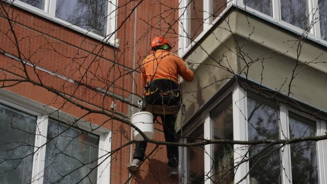 Dirty industrial climber in orange clothes working near buildings outside. Worker hanging on ropes and painting the wall with sealant. House renovation