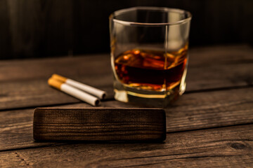 Glass of brandy with cigarettes and the empty wooden plank on an old wooden table. Angle view, focus on the wooden plank