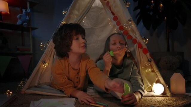 Children in cute decorative makeshift hut blowing bubbless at home in evening. Girl and boy lying on floor while spending good time. Concept of leisure and careless childhood.