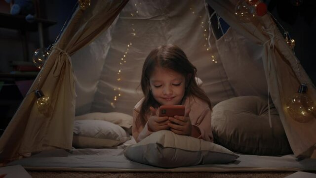 Adorable small girl spending time in decorative tent with lightings in evening playroom. Cute little kid lying on floor and smiling while watching cartoon on smartphone. Zoom in.