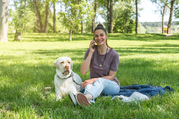 Fototapeta premium Young beautiful woman talking on her smartphone while is playing and enjoying with her pet dog the golden Labrador Retriever in the park on a sunny and beautiful day