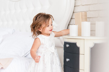 Cute little girl stands near the dresser. She is wearing a simple white dress. Bright room. European race.
