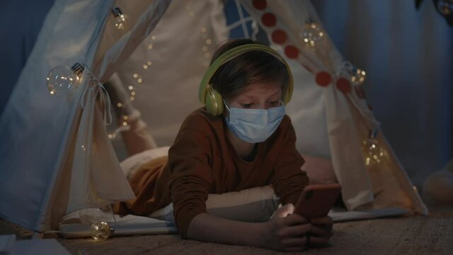 Young boy with headphones spending time in decorative makeshift hut at home in evening. Teen in medical mask lying on floor while using and scrolling smartphone.Concept of leisure.