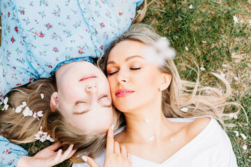 Mom and daughter are lying on the grass and falling tree flowers, blonde hair, park or forest