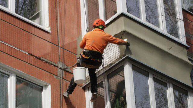 Dirty industrial climber in orange clothes working near buildings outside. Worker hanging on ropes and painting the wall with sealant. House renovation