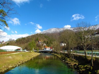 Idrijca river flowing through Idrija in Littoral region of Slovenia with forest covered hills above and the top covered in snow