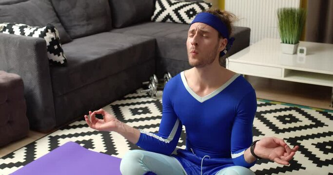 Comical Caucasian Man calms down practicing Yoga breathing in Lotus Pose. Positive Attractive Guy Meditating and looking at camera with a Funny Face, Grimacing. Wears Retro-Style. Meditation at home.