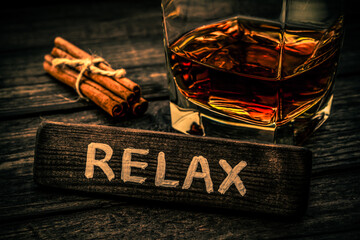 Glass of brandy with cinnamon sticks tied with jute rope and the wooden plank on it is an inscription "RELAX" on an old wooden table. Close up view, focus on the inscription