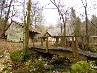 Old abandoned traditional house on a meadow in Gorenjska, Slovenia with stream wooden bridge crossing a stream and a collapsing wooden shack in front