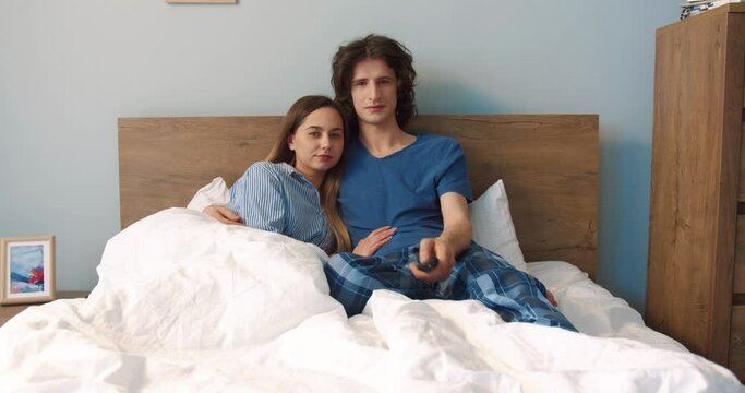 Full length view of the couple of the man and woman embracing at the cozy bed and watching movie together at the weekend. Family relationships and pastime concept