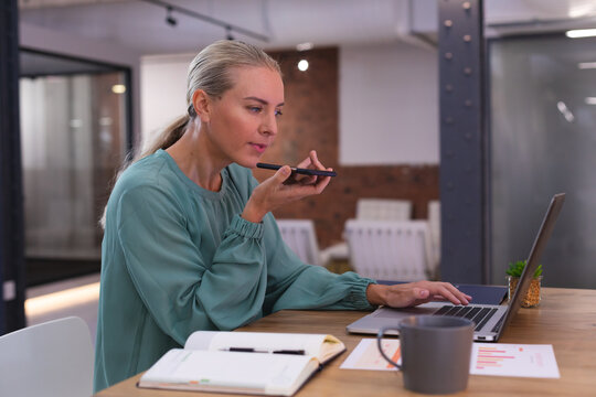Caucasian woman talking on smartphone while using laptop at modern office