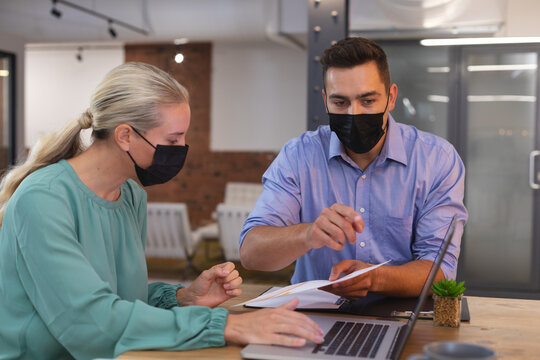 Caucasian male and female office colleagues wearing face masks discussing over a document at office