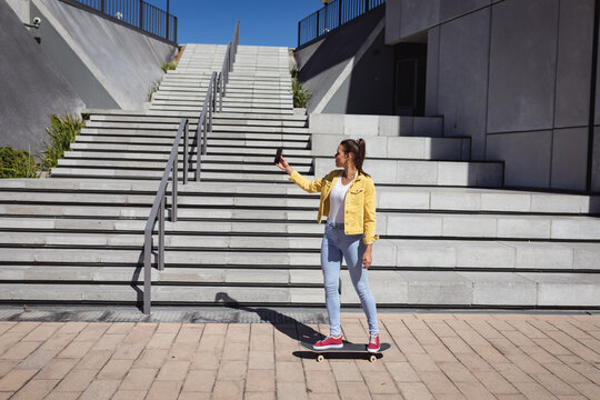 Smiling caucasian woman standing on skateboard and taking selfie next to stairs