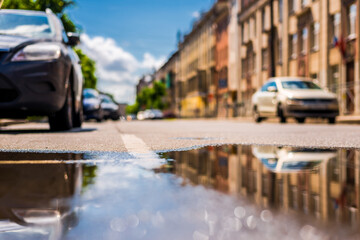 Sunny day after the rain in the city, parked on the street cars on the background of the facades of the house. Close up view from the level of the puddle on the pavement