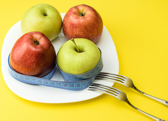 Red and yellow apples, wrapped with a measuring tape, on a white plate, on a yellow background, next to two table forks. The concept of healthy food, fitness, diet, weight loss. Selective focus.