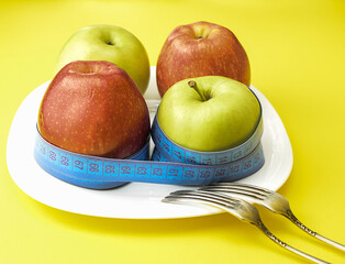 Red and yellow apples, wrapped with a centimeter tape on a white plate, on a yellow background, next to two table forks. The concept of healthy food, fitness, diet, weight loss. Selective focus.