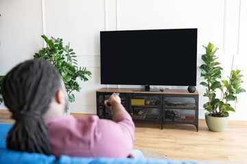 Back view African-American guy with dreadlocks holds remote controller and switching tv channels,...