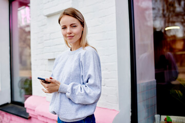 Half length portrait of young Caucaisan tourist in casual grey pullover looking at camera during vacations, attractive hipster girl with modern smartphone technology for blogging posing outdoors