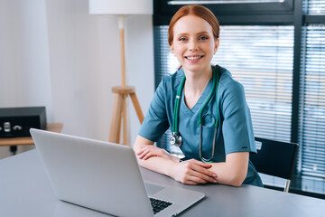 Portrait of attractive smiling young female doctor in blue green medical uniform sitting at desk...